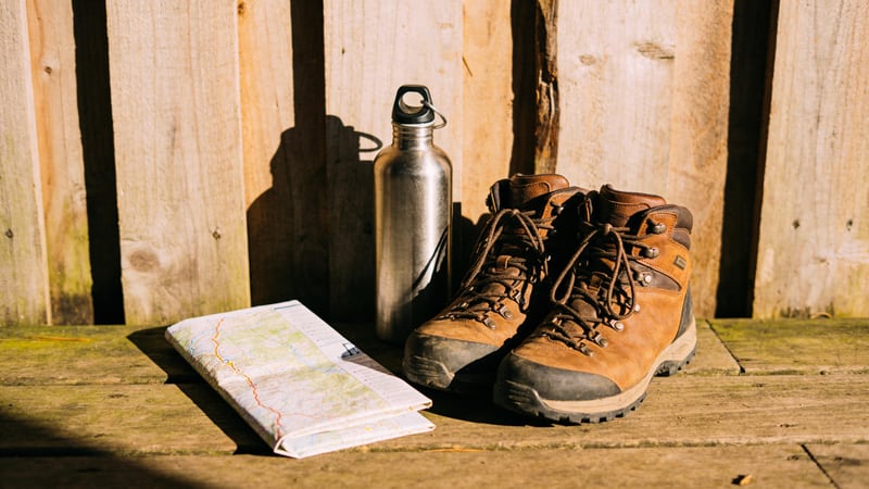 Wanderschuhe und Outdoor-Karte liegen auf rustikalem Holzboden, eine Trinkflasche daneben, warmes Morgenlicht — Aktiv-Leben mit der Barmenia Unfallversicherung